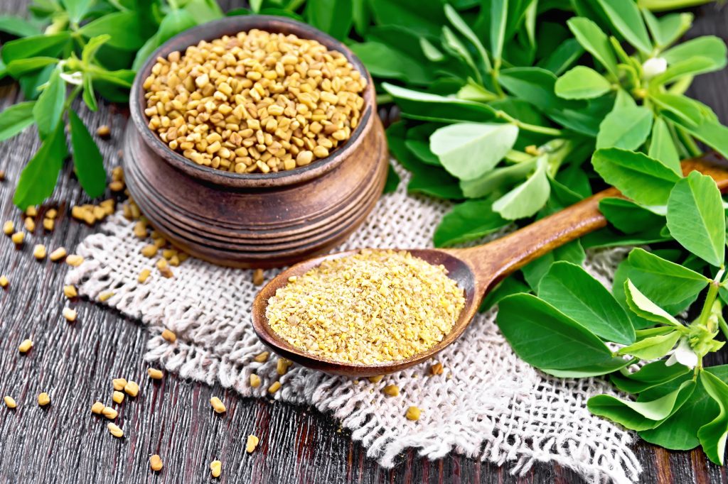 Ground fenugreek in a spoon and seeds in a bowl on a burlap napkin with leaves on wooden board background