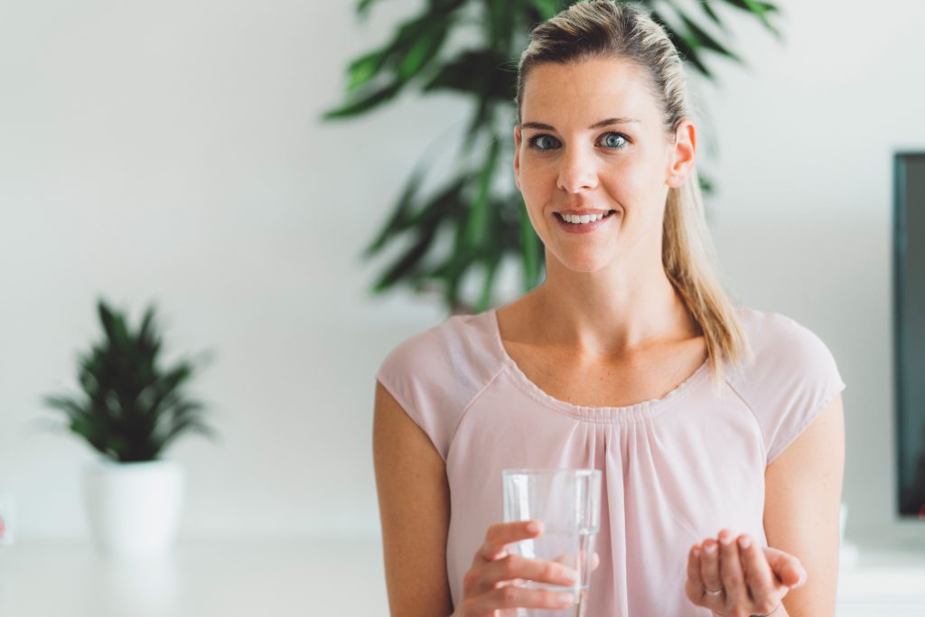 Smiling young caucasian woman taking her morning supplements, vitamin pills with a glass of water at home. High quality photo