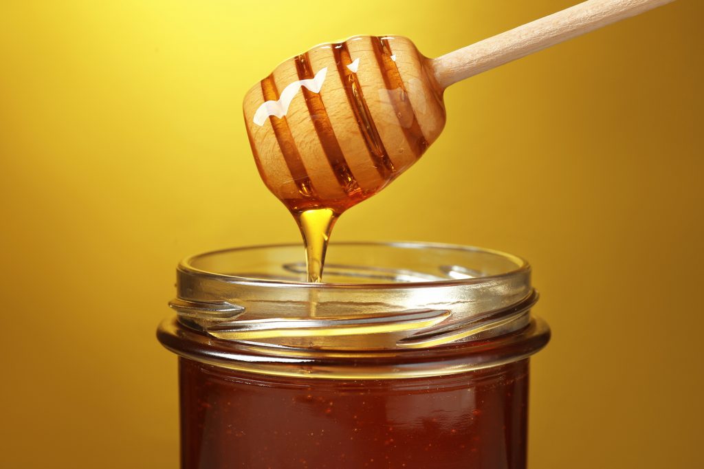 Pouring tasty honey from dipper into glass jar on golden background, closeup